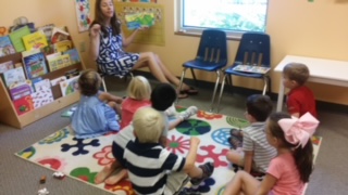 A woman reads to children sitting on a colorful rug in a classroom with bookshelves and chairs nearby.