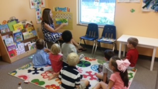 Young girl standing and reading to a group of children sitting on a colorful rug in a classroom.