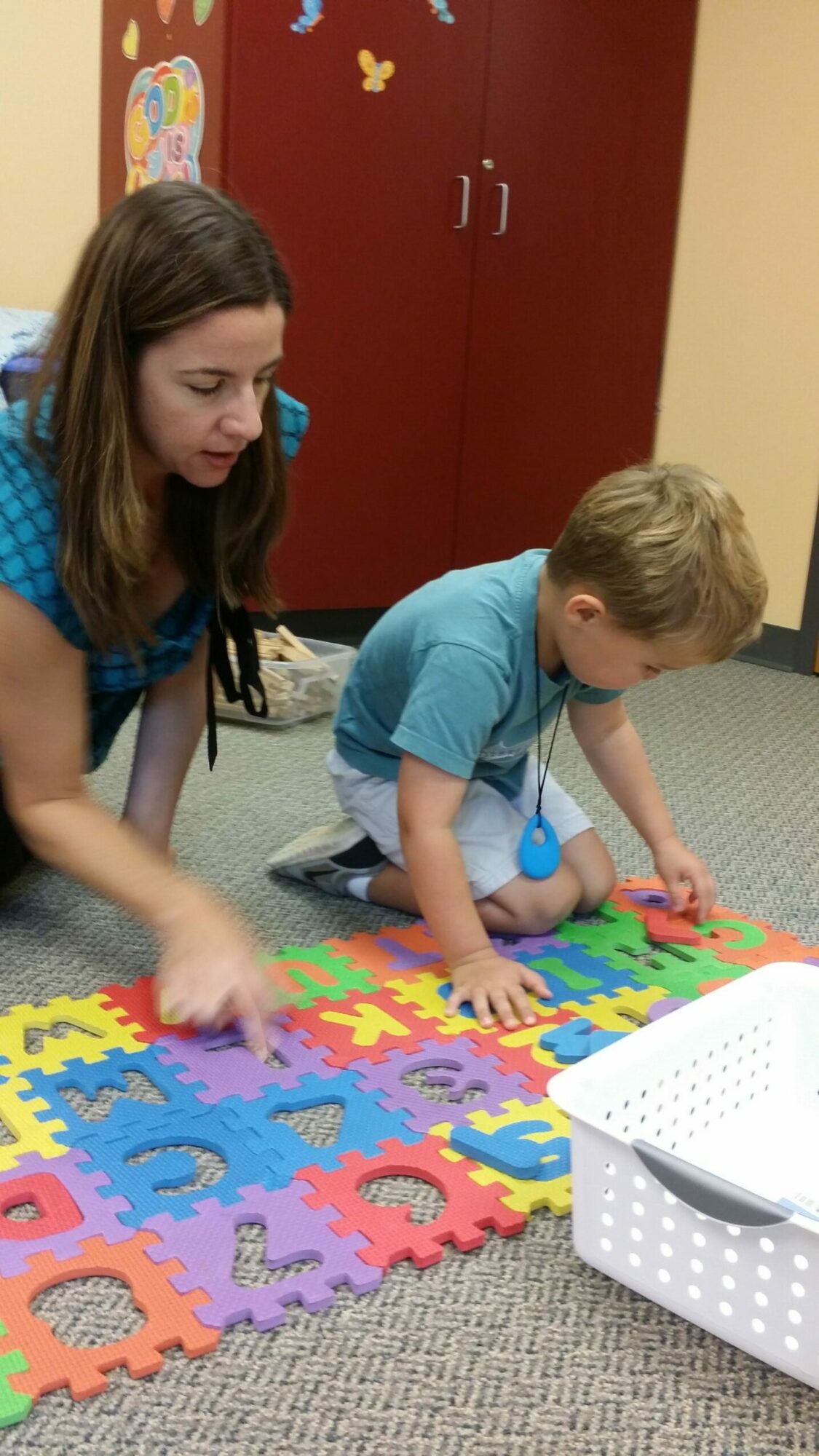 Child and woman playing with colorful puzzle pieces on the floor in a room with a red cabinet.