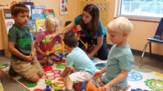A woman interacts with four children on a colorful rug in a classroom, engaging in a group activity.
