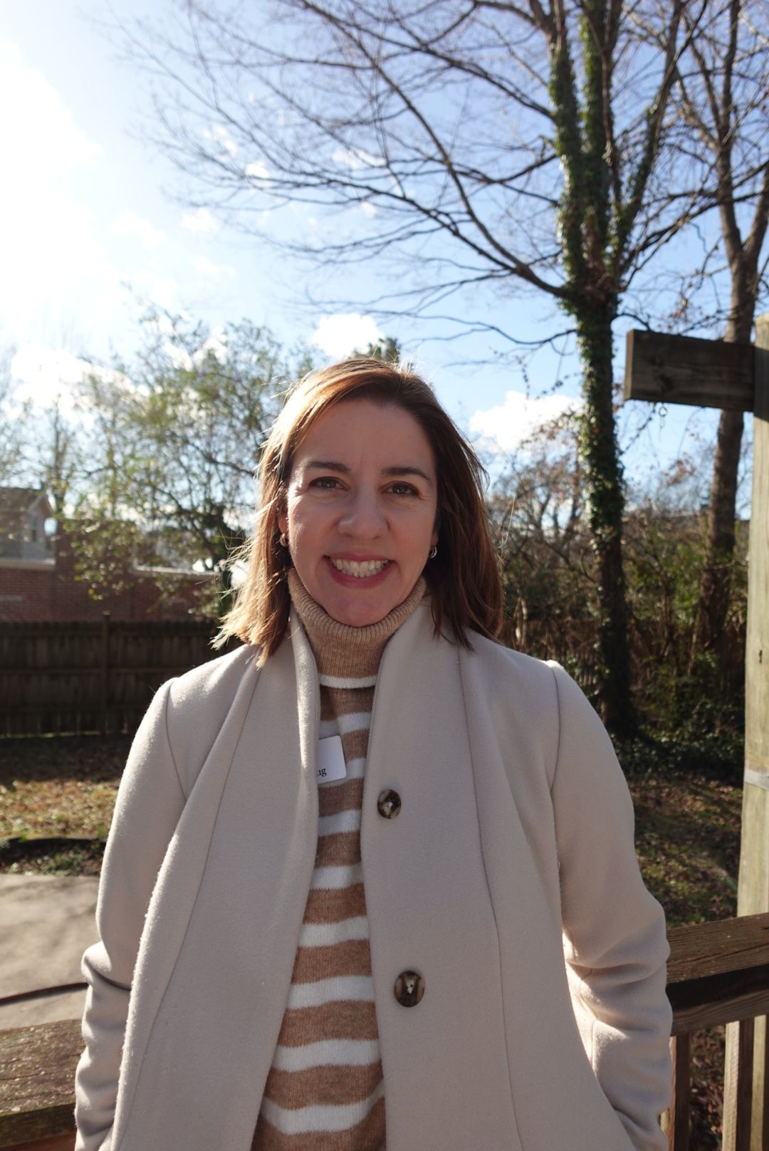 Woman smiling outdoors with trees and a wooden structure in the background.
